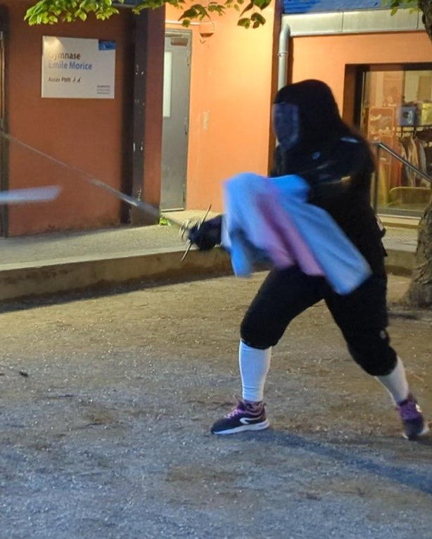 Photo de deux personnes en équipement de protection noir, équipées de rapières, en plein duel. La personne de gauche a une dague dans sa main gauche, et la personne de droite tient une cape enroulée autour de sa main, et est sur le point de la projeter en