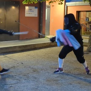 Photo de deux personnes en équipement de protection noir, équipées de rapières, en plein duel. La personne de gauche a une dague dans sa main gauche, et la personne de droite tient une cape enroulée autour de sa main, et est sur le point de la projeter en